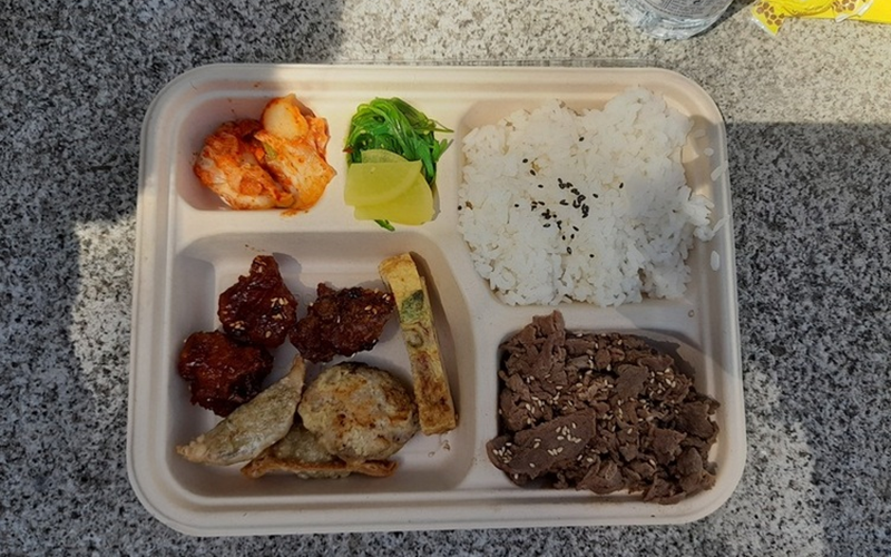 La foto muestra el dosirak ofrecido en la degustación de gastronomía coreana, llevada a cabo el 11 de mayo en los jardines de la Facultad de Derecho de la Universidad Complutense de Madrid, España. De izquierda a derecha, según las manecillas del reloj: kimchi (col fermentada), alga wakame, nabo encurtido, arroz blanco cocido con semillas de sésamo negro encima, dakgangjeong (pollo frito al estilo coreano con salsa picante), gyeranmari (tortilla de huevo con diferente variedad de verduras), mandu (empanadillas coreanas), wanjajeon (bolitas de carne picada aplanadas) y bulgogi (carne de ternera marinada en una salsa dulce) con semillas de sésamo blanco encima. 