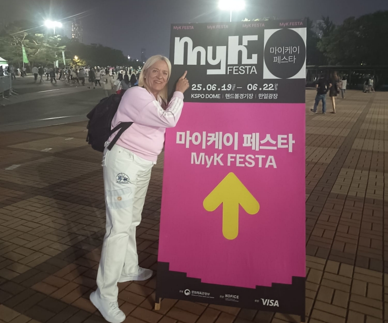 La reportera honoraria Claudia Möller posa junto a un cartel informativo del MyK FESTA, en las afueras del KSPO Dome en Seúl. | Claudia García Bueno